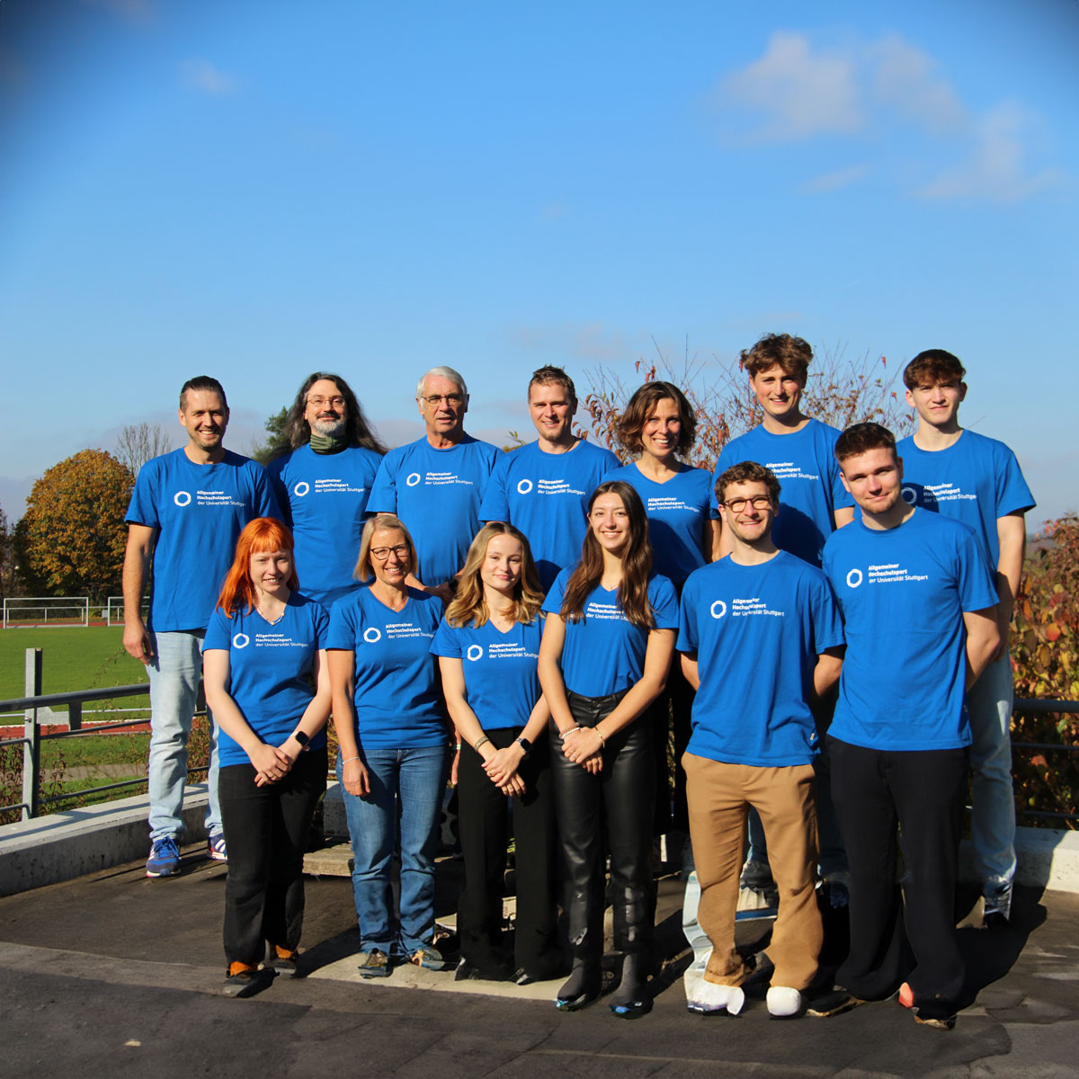 Das Bild zeigt 13 lachende Mitglieder des Allgemeinen Hochschulsports in ihren blauen Team T-Shirts. Sie stehen in zwei Reihen vor einem Sportplatz und klarem Himmel.