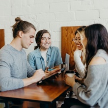 Eine Gruppe mit drei Frauen und einem Mann sitzt sich an einem Tisch gegenüber. Sie unterhalten sich, während Laptops aufgeklappt sind und Kaffeetassen herumstehen. Es könnte eine Lerngruppe sein.