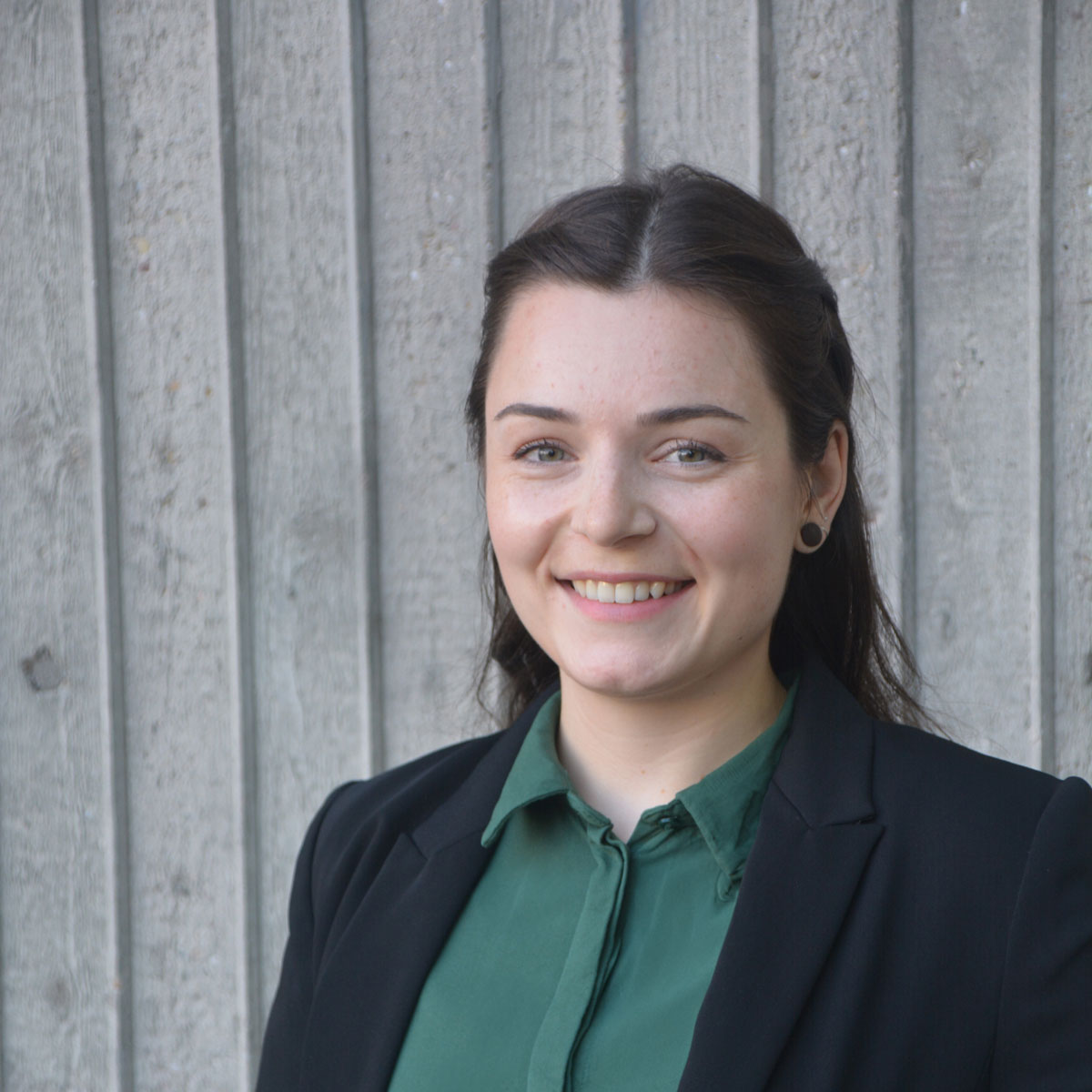Svenja hat sehr dunkle, lange Haare mit Mittelscheitel, grüne Augen und helle Haut. Sie trägt eine grüne Bluse und einen schwarzen Blaizer. Sie lächelt sympathisch.