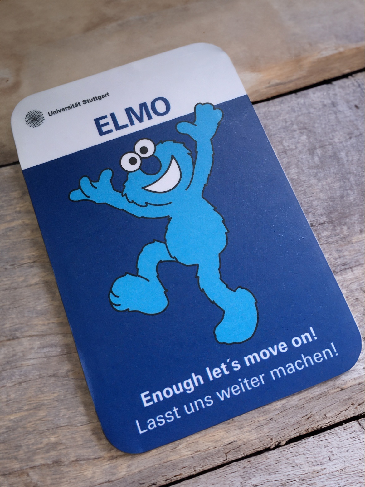 Eine Karte mit einer blauen plüschigen Comicfigur wird in eine Kamera gehalten. Dabei steht der Schriftzug „ELMO - Enough, let's move on!“