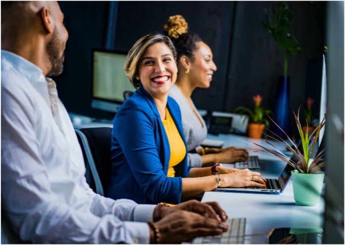 A diverse group of professionals sitting at a desk with laptops, smiling and engaging with each other in a collaborative work environment. The woman in the foreground, dressed in a blue blazer and yellow top, is looking directly at the camera with a bright smile, while her colleagues work alongside her.
