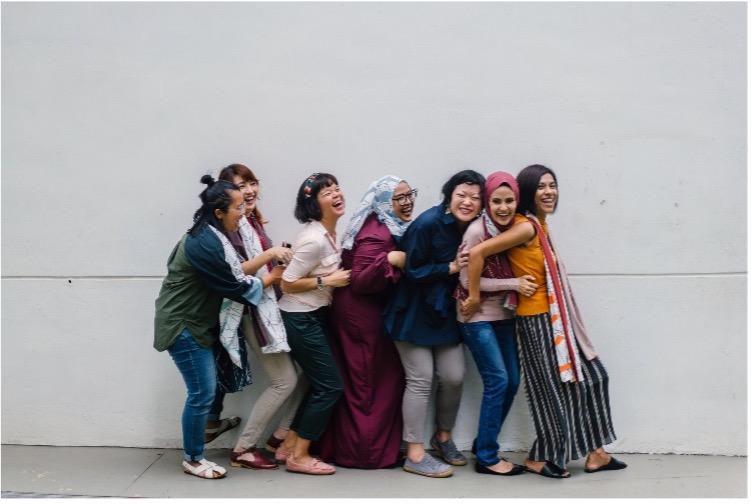 A group of women of diverse ethnic backgrounds standing in a playful line, holding onto each other and laughing. They are casually dressed in a variety of styles, including headscarves and colorful clothing, posing against a simple white wall. The atmosphere is joyful and relaxed, emphasizing camaraderie and friendship