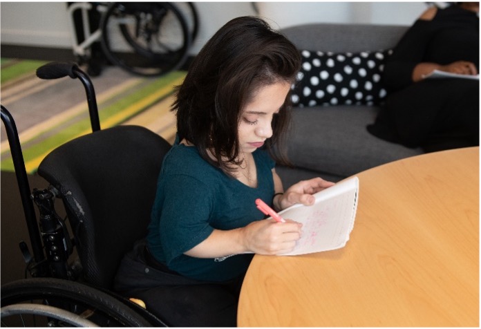 A woman seated in a wheelchair writes in a notebook with a pink pen at a round table. She is focused on her writing, with a calm and engaged expression. In the background, part of another person is visible, seated on a couch. The setting appears to be an informal, collaborative space, emphasizing participation and focus.